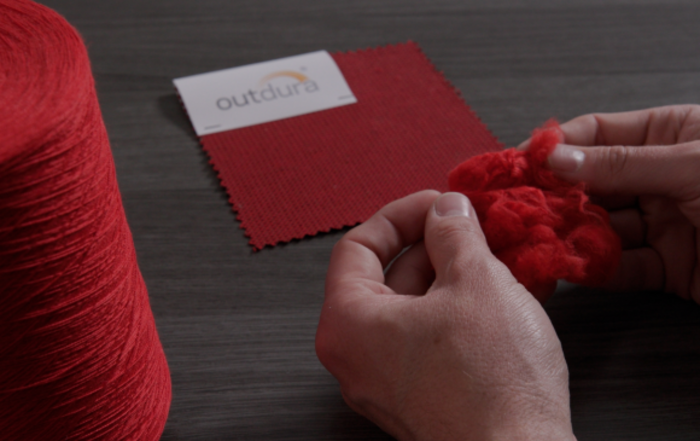 A set of hands holding red fibers in front of a sample of Outdura fabric